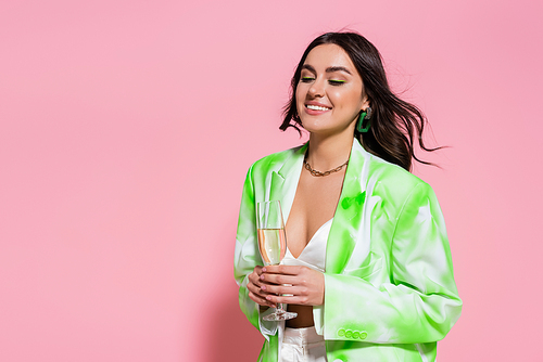 Positive woman in necklace and jacket holding glass of champagne on pink background