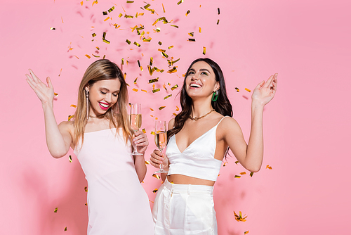 Pretty friends holding champagne under falling confetti on pink background