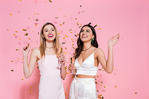 Positive brunette woman with champagne standing near friend and confetti on pink background