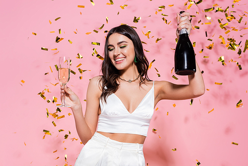 Pretty brunette woman holding bottle and glass of champagne under golden confetti on pink background