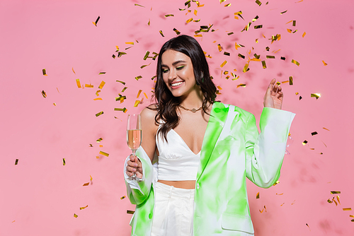 Smiling woman looking at glass of champagne under confetti on pink background