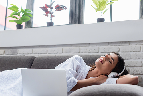 peaceful young adult woman with closed eyes in headphones resting on couch near laptop at home