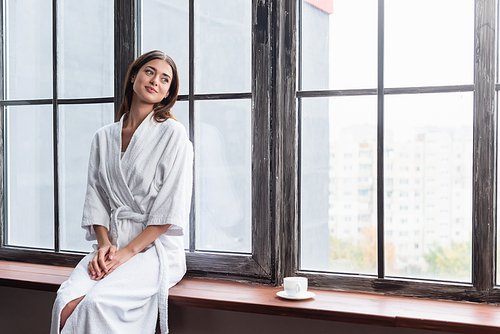 pretty young adult woman in bathrobe sitting on sill with cup of coffee near window at home