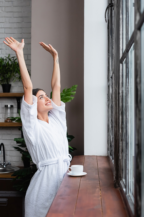 happy young adult woman in bathrobe standing near window with hands in air in modern living room