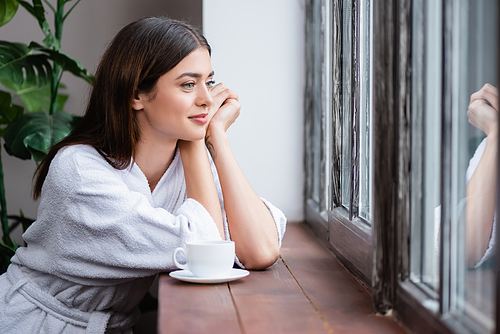 smiling young adult woman sitting with hands near face and looking out window at home