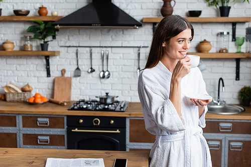 smiling young adult woman in bathrobe drinking coffee near table with newspaper and cellphone in modern loft
