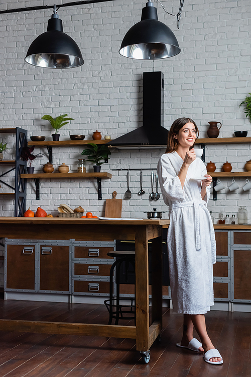 full length view of happy young adult woman in bathrobe drinking coffee in modern loft