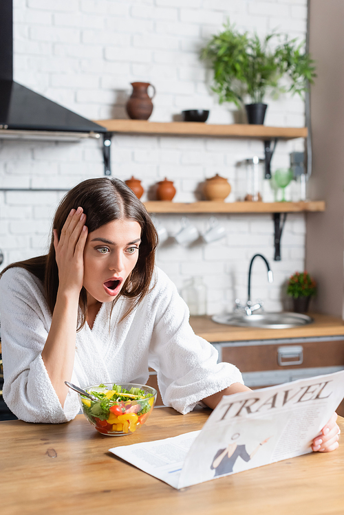 excited young adult woman in bathrobe eating salad and reading travel newspaper in modern kitchen