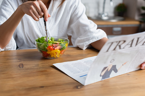 partial view of young adult woman in bathrobe eating salad and reading travel newspaper in kitchen