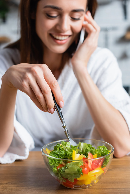 smiling young adult woman in bathrobe eating salad and speaking on cellphone in kitchen