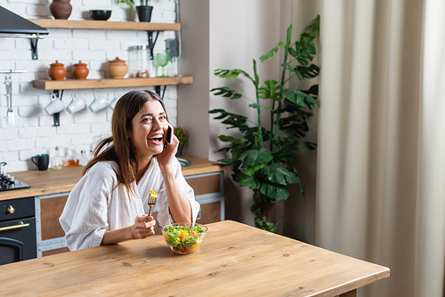young adult woman in bathrobe eating salad and speaking on cellphone in modern kitchen