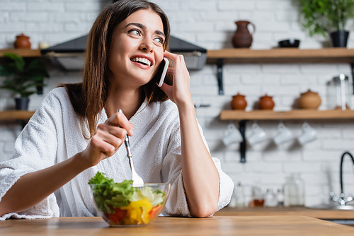 brunette young adult woman in bathrobe eating vegetables salad and speaking on cellphone in modern kitchen