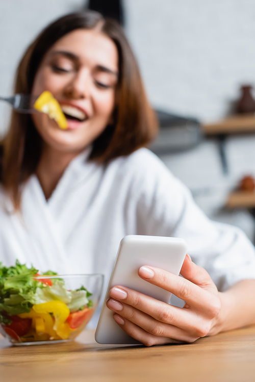 cheerful young adult woman in bathrobe eating salad and using cellphone in modern kitchen, focus on foreground