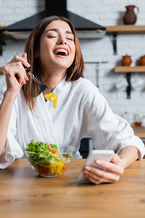 laughing young adult woman in bathrobe eating vegetables salad and using cellphone in modern kitchen