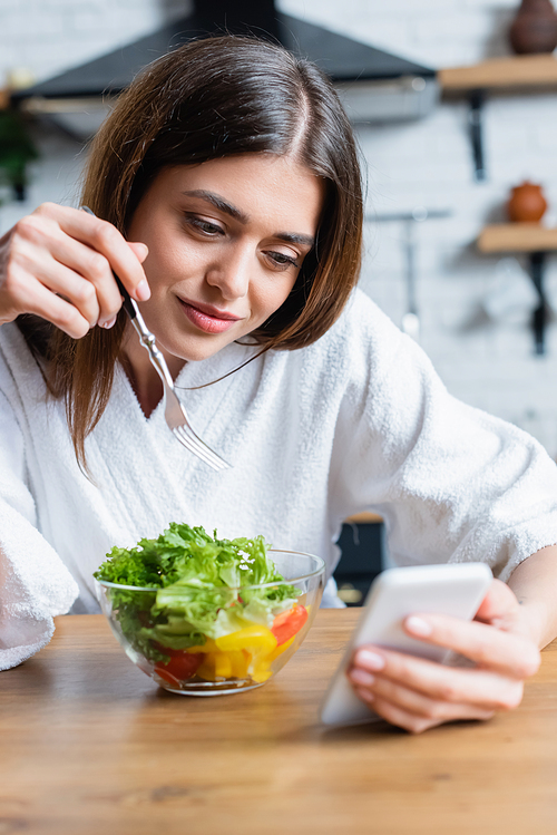 smiling young adult woman in bathrobe eating vegetables salad and looking at cellphone in modern kitchen