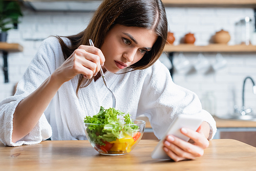upset young adult woman in bathrobe eating vegetables salad and using cellphone in modern kitchen