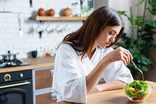 sad young adult woman in bathrobe eating vegetables salad in modern kitchen