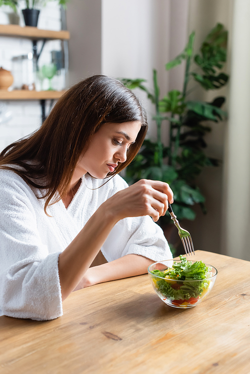 disappointed young adult woman in bathrobe eating vegetables salad in modern kitchen