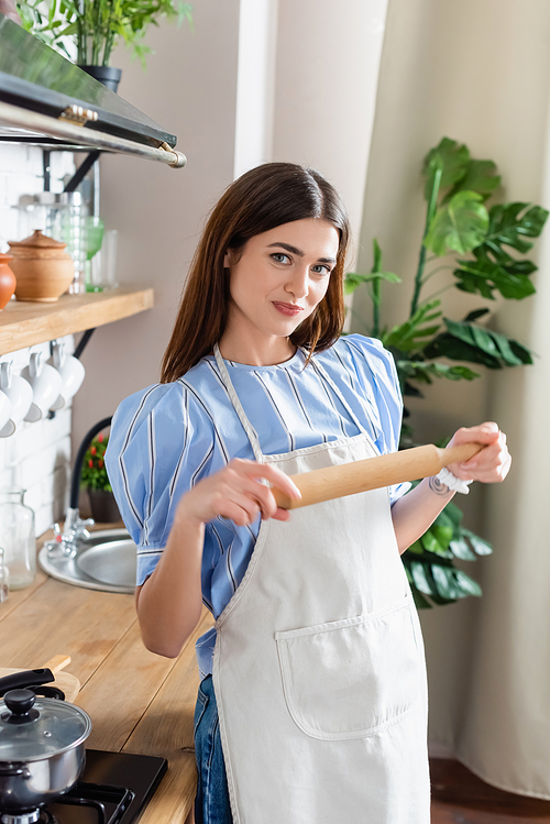 pretty young adult woman in apron standing with dough rolling pin in modern kitchen