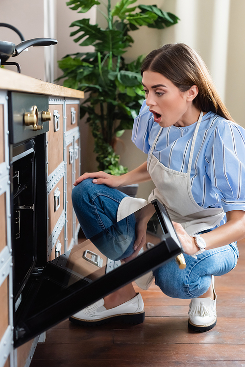 astonished young adult woman in apron looking into oven with open mouth in modern kitchen