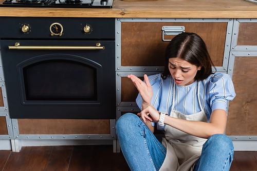 confused young adult woman in apron sitting near oven and looking at wristwatch in modern kitchen