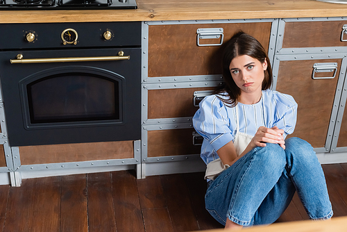 tired young adult woman sitting on floor near oven with in modern kitchen
