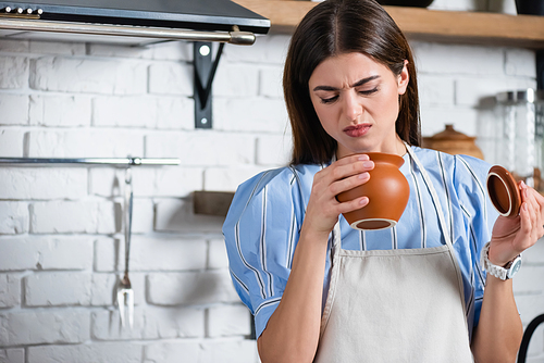 young adult woman in apron looking at clay pot with disgust in kitchen