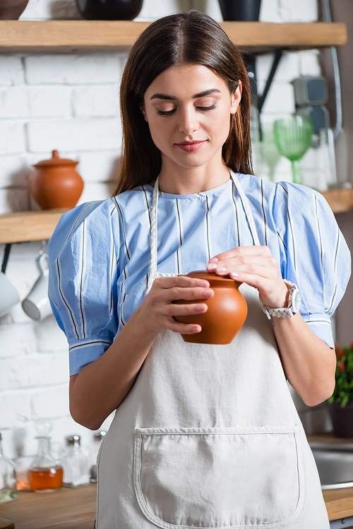 cheerful young adult woman in apron holding clay pot in hands in kitchen