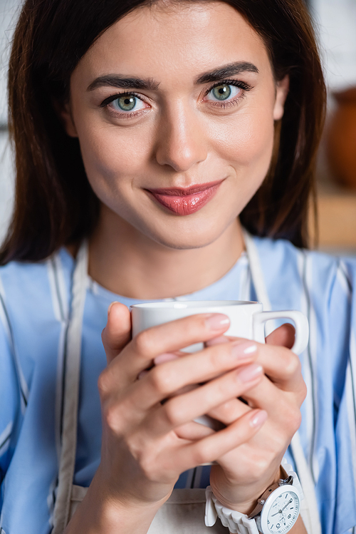 portrait of smiling young adult woman holding cup of coffee in hands on blurred background