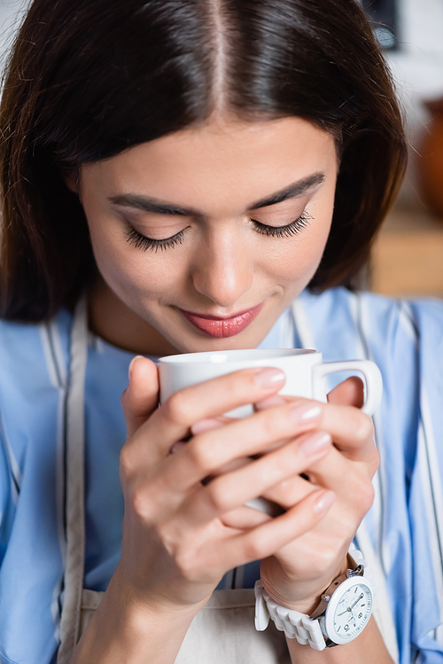 portrait of smiling young adult woman smelling aroma of coffee in white cup on blurred background