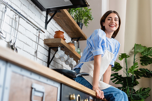 low angle view of smiling young adult woman in apron sitting in modern kitchen