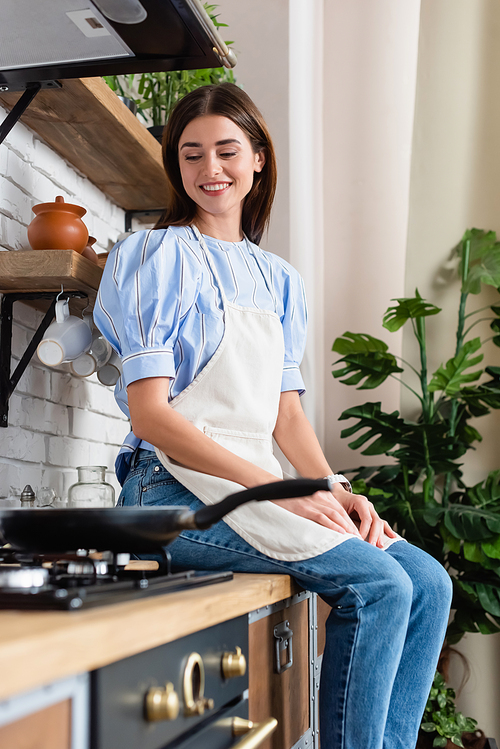 smiling young adult woman in apron sitting near frying pan in modern kitchen