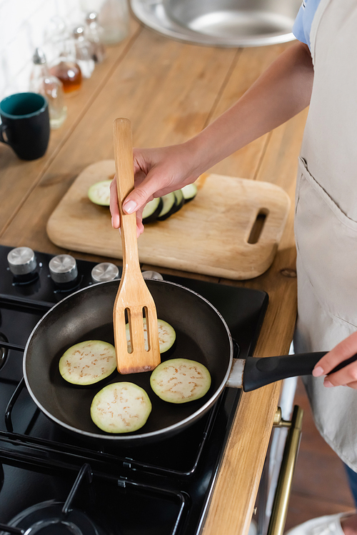 cropped view of young adult woman cooking slices of eggplant in frying pan in kitchen