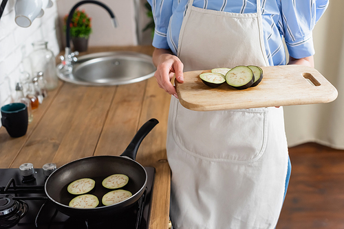 cropped view of young adult woman holding slices of eggplant on cutting board near frying pan in kitchen