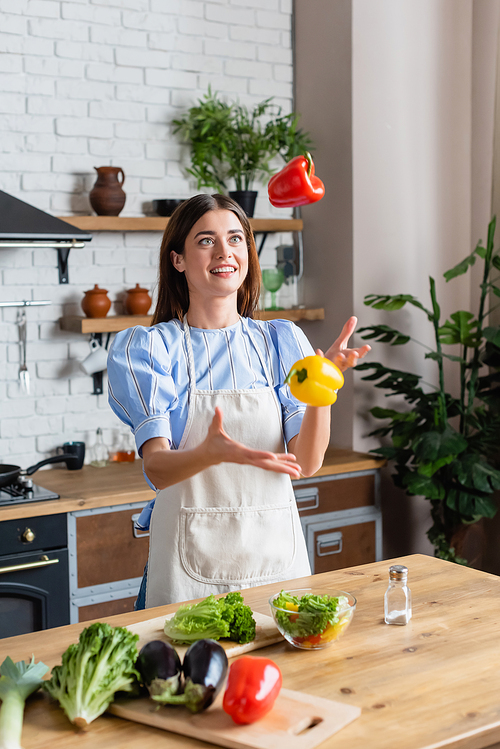 smiling young adult woman juggling with fresh peppers in kitchen