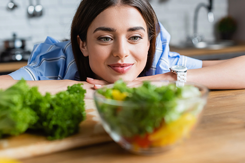 smiling young adult woman looking at prepared vegetables salad in kitchen