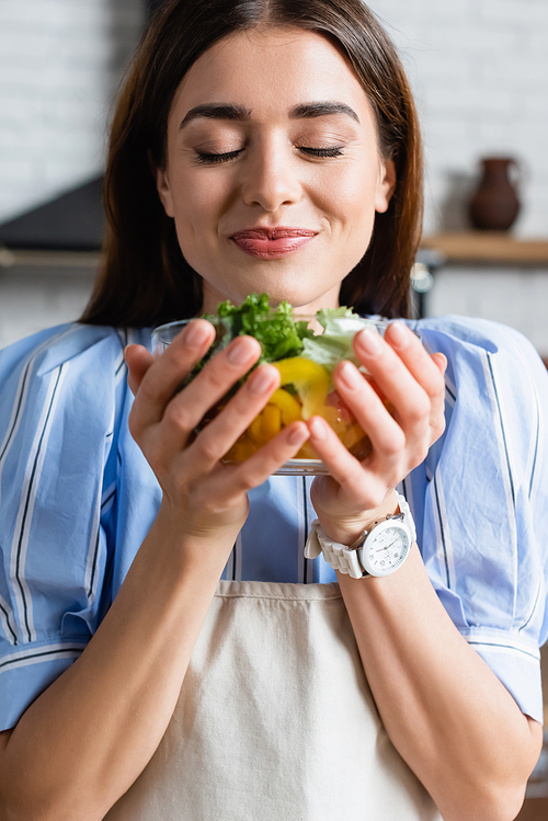 smiling young adult woman enjoying smell of fresh vegetables salad with closed eyes in kitchen