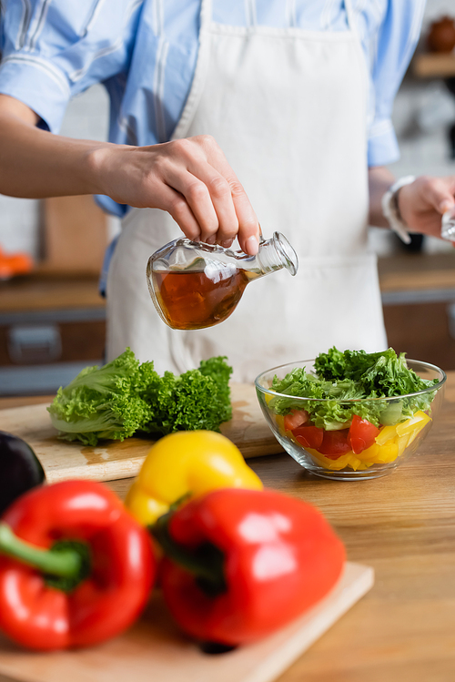 partial view of young adult woman seasoning fresh vegetables salad with olive oil in kitchen