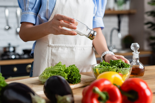 partial view of young adult woman in apron seasoning fresh vegetables salad with salt mill in kitchen