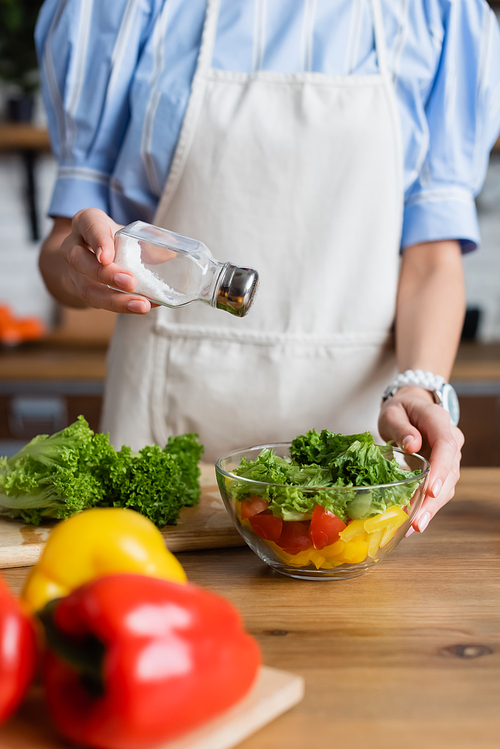 partial view of young adult woman in apron seasoning fresh vegetables salad with salt mill in modern kitchen