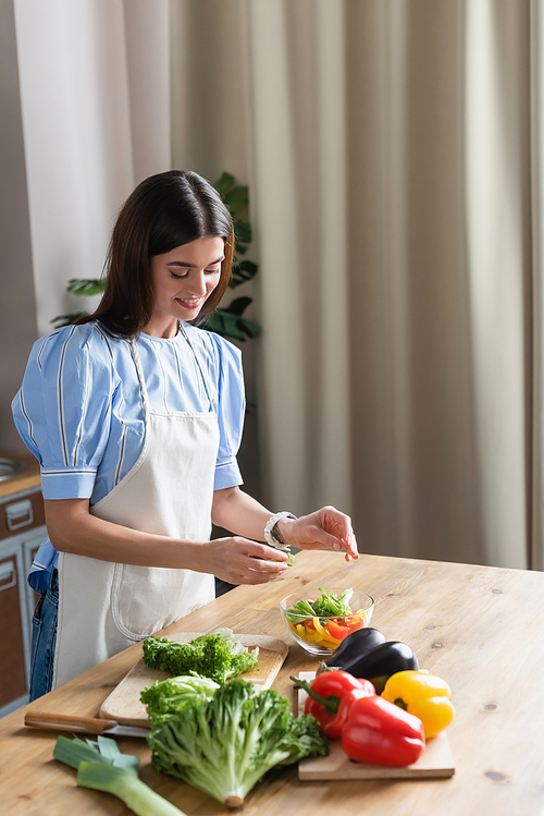 smiling young adult woman in apron cooking fresh vegetables salad in kitchen