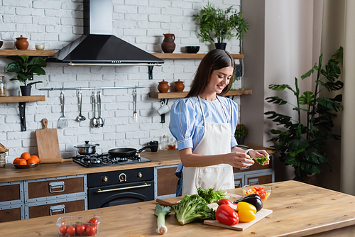 happy young adult woman in apron preparing vegetables salad in modern kitchen