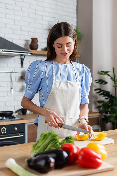 smiling young adult woman chopping yellow pepper on cutting board in kitchen