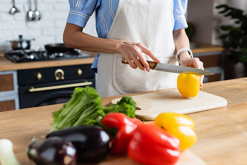 partial view of young adult woman cutting yellow pepper on cutting board in kitchen