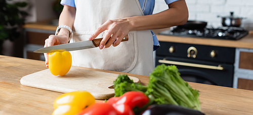 partial view of young adult woman cutting yellow pepper on cutting board in kitchen, banner