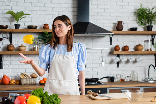 attractive young adult woman throwing yellow pepper into air in kitchen
