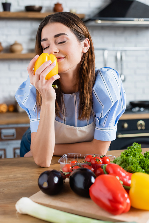 smiling young adult woman smelling fresh yellow pepper in kitchen