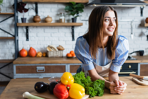 smiling young adult woman standing near table with vegetables in kitchen