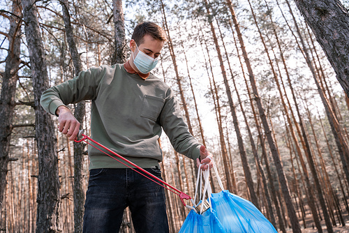 volunteer in medical mask holding pick up tool near rubbish in forest