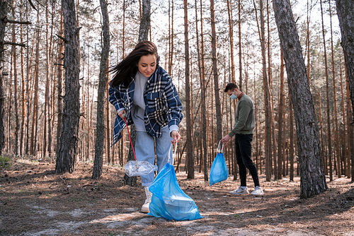 young woman and man with trash bags picking up rubbish with grabber tools in forest